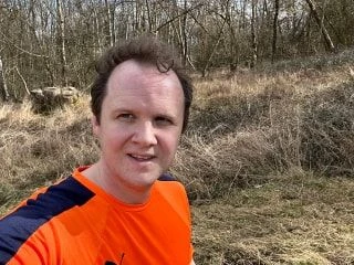 Man wearing a bright orange running shirt taking a selfie on a trail surrounded by dry grass and leafless trees.