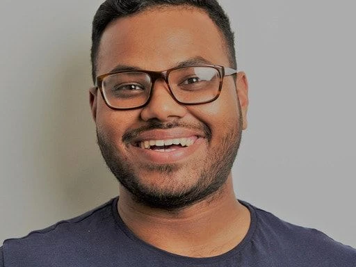 Man smiling broadly wearing glasses in front of a grey background.