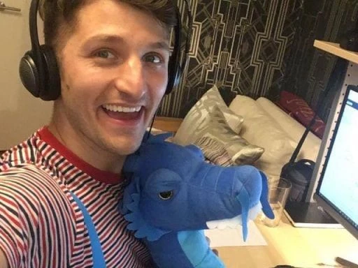 Man smiling broadly wearing headphones sitting at a computer desk in front of a screen. He is holding a bright blue dragon plush toy.