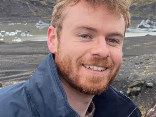 Smiling man with reddish-brown hair and beard, outdoors with rocky terrain and a lake with ice in the background.