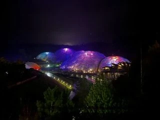 Colourfully lit Eden Project biodomes glowing at night surrounded by trees.