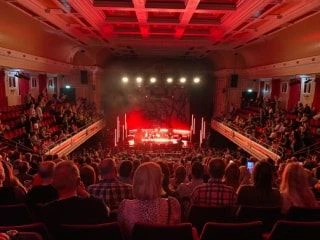 Audience seated inside a theatre hall enjoying a live performance on a brightly lit stage.